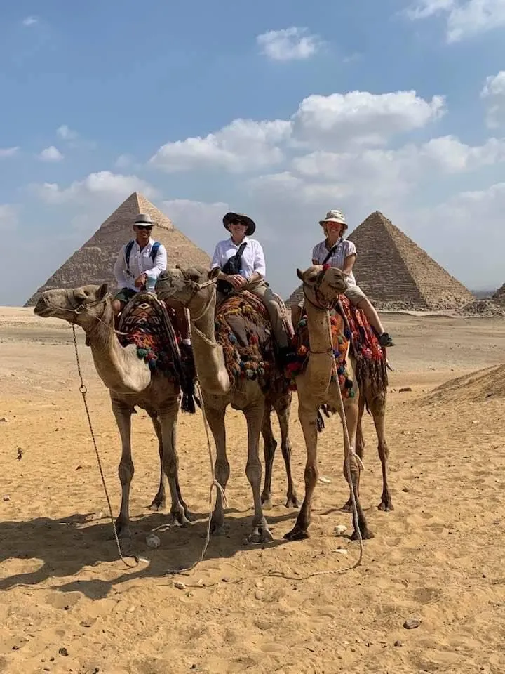 three people on camels in front of pyramids